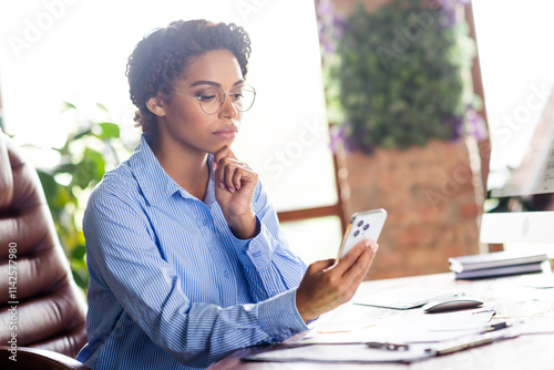 Young businesswoman in office setting using smartphone with focused expression, wearing elegant blue blouse
