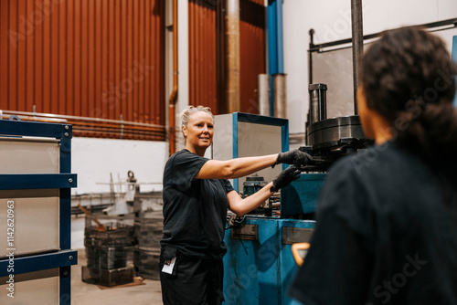 Female worker wearing gloves while working on machine and talking with female colleague in metal manufacturing factory photo