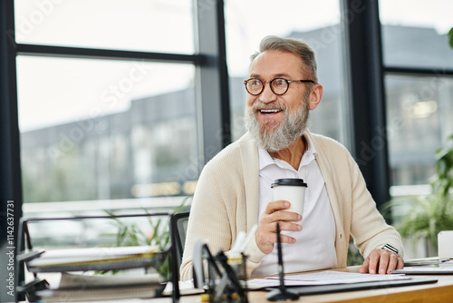 Handsome senior man smiles while holding a coffee cup at his workspace in a bright office.