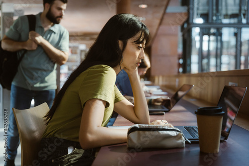 Focused young woman studying on laptop while leaning on elbow at desk in university library photo