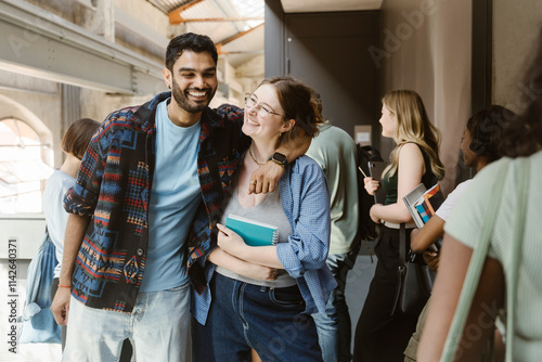 Happy man standing with arm around female friend at college corridor photo