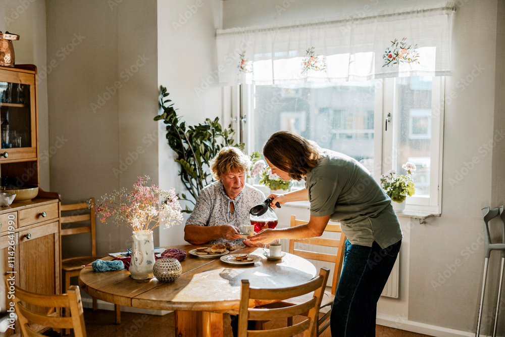 Female healthcare worker serving coffee to elderly woman sitting at dining table Female healthcare worker serving coffee to elderly woman sitting at dining table