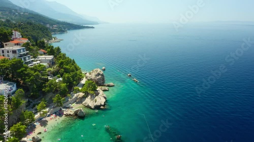 Hotels and houses on the mountainside on the seafront. Tourists sunbathe on the beach, swim in the sea, and ride paddle boards. A seaside tourist town on the picturesque Adriatic coast of Croatia.