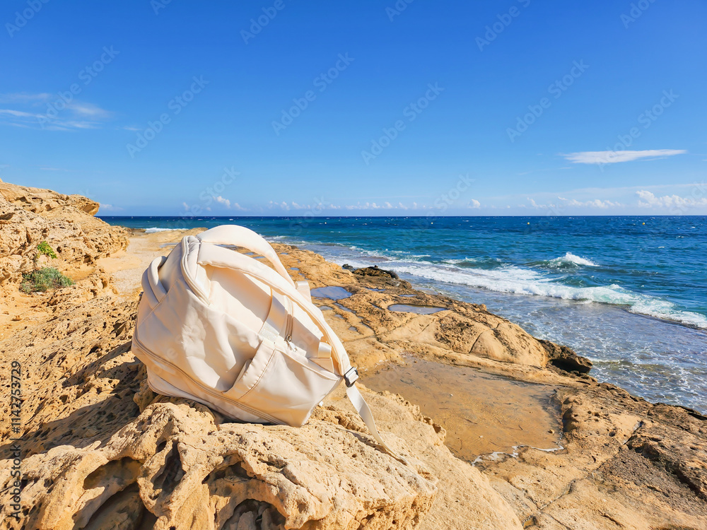 White backpack resting on rugged rocks with a coastal background, blue sea, and waves under sunlight. White backpack resting on rugged rocks with a coastal background, blue sea, and waves under sunlight.