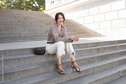 Woman in silk striped brown shirt, white denim pants and black heeled sandals sitting on the stairs outside on a city street. Trendy elegant casual outfit. Street fashion.