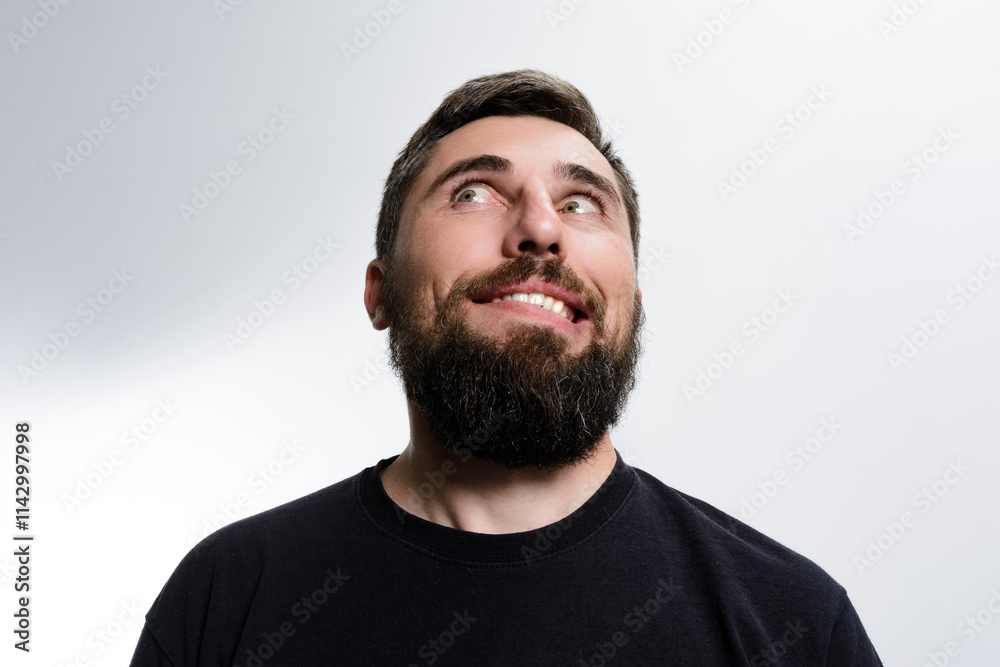 A man with a beard looks upwards with a curious and slightly mischievous expression on his face against a plain background. A man with a beard looks upwards with a curious and slightly mischievous expression on his face against a plain background.