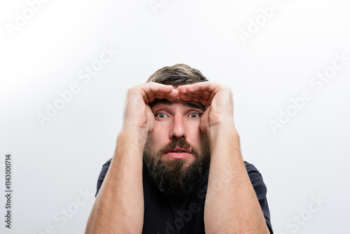 A bearded man against a white background holds his hands around his eyes, peering through them with a look of surprise and curiosity. photo