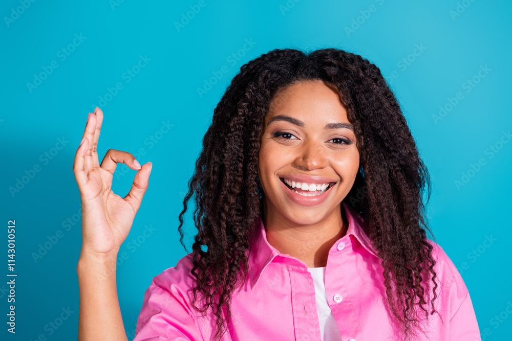 Charming young woman in a pink shirt gives a positive sign against a bright blue background, showcasing her lovely curls Charming young woman in a pink shirt gives a positive sign against a bright blue background, showcasing her lovely curls