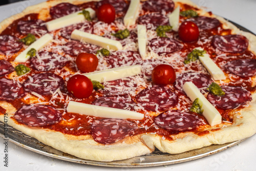 Pizza is prepared for baking. Raw pepperoni pizza with sausage, mozzarella cheese, cherry tomatoes and basil on a baking wheel