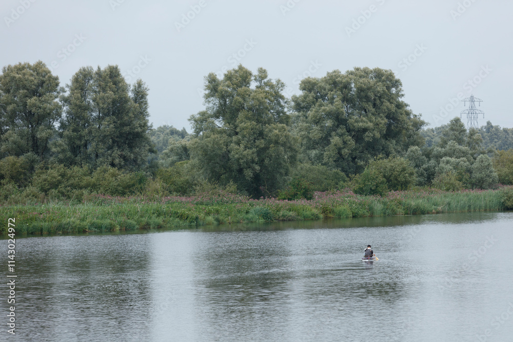 Calm river scene with a lone kayaker, surrounded by trees and vegetation. Gray sky, overcast day. Calm river scene with a lone kayaker, surrounded by trees and vegetation. Gray sky, overcast day.