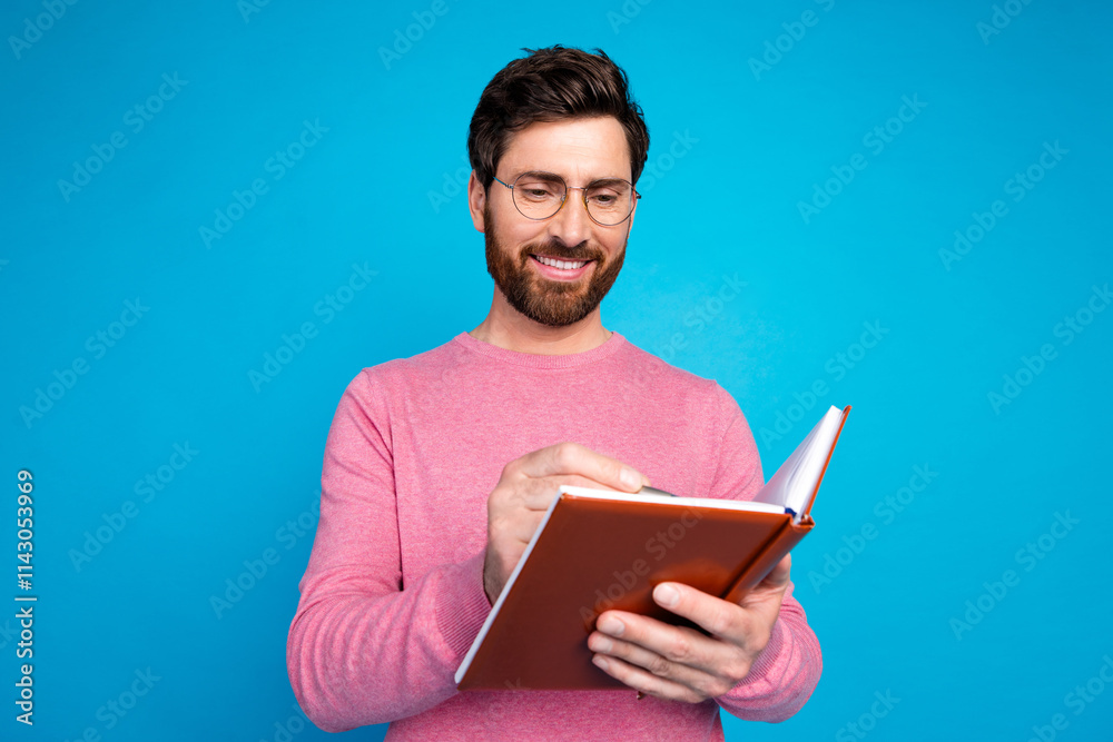 Smiling man with a beard in casual pink sweater holding a notebook, writing against a vibrant blue background Smiling man with a beard in casual pink sweater holding a notebook, writing against a vibrant blue background