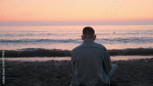 Rear view of Caucasian man sitting alone on black sand beach on the coast and enjoying beautiful sunset over the sea. Thoughtful male traveler enjoy freedom and tranquility of contemplating nature