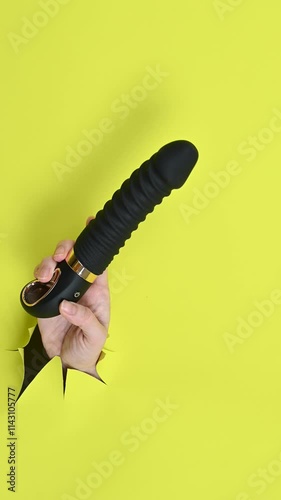Female hand with black sex toy sticking out from yellow paper background. Copy space.