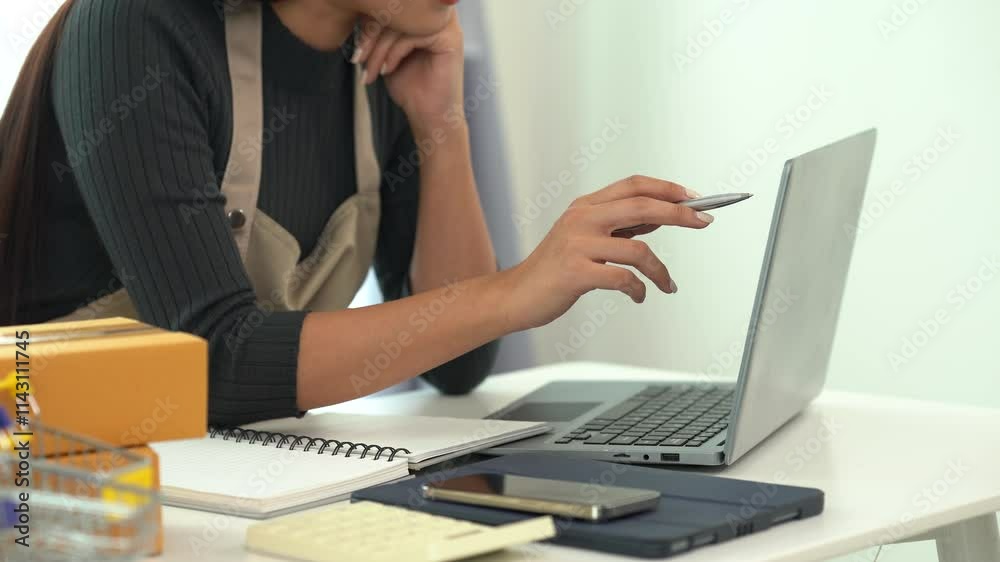 Focused Online Business Owner: A female entrepreneur thoughtfully works on her laptop, surrounded by packaging materials and small business tools.  The image portrays dedication and concentration. 