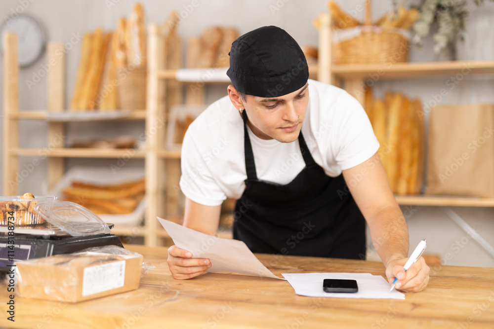 Young baker reading documents and writing notes on paper in the interior of a private bakery Young baker reading documents and writing notes on paper in the interior of a private bakery