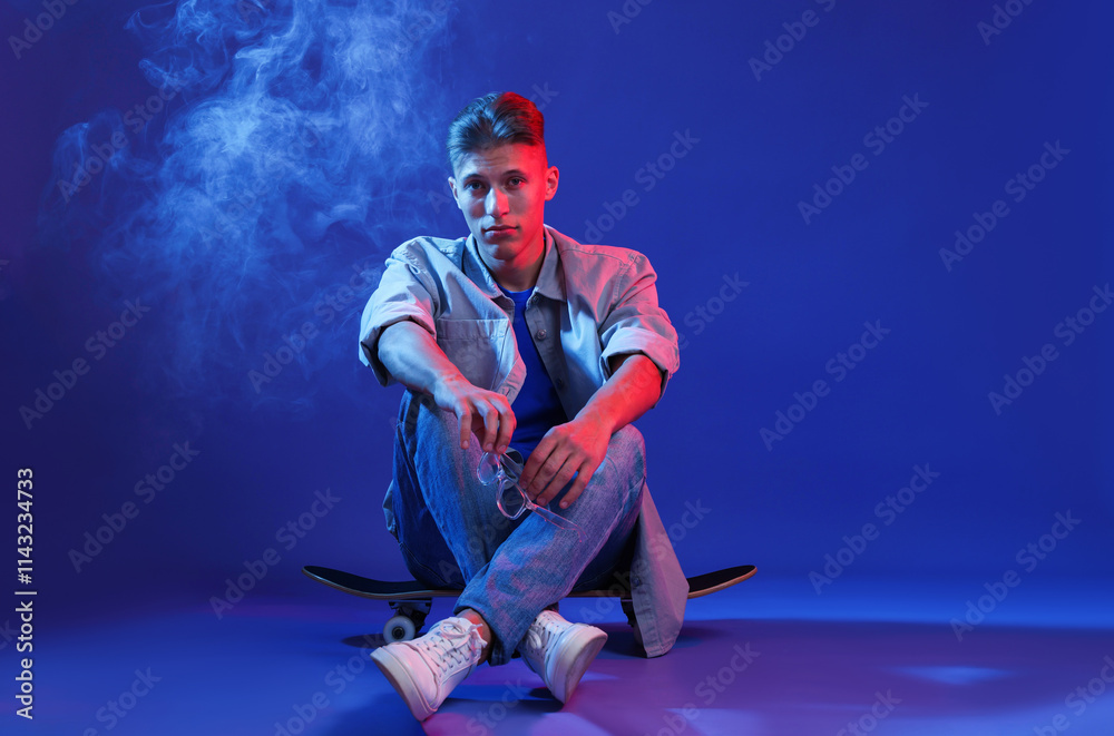 Fototapeta premium Handsome man sitting on skateboard in smoke and color lights against blue background