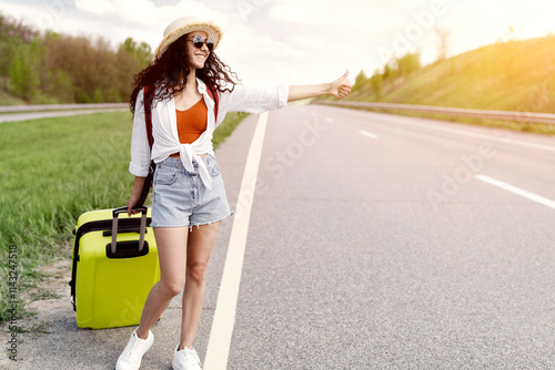 Full length of happy young woman with backpack and suitcase trying to stop car, showing thumb up, asking for free ride, hitchhiking on roadside, copy space. Traveling by autostop concept photo