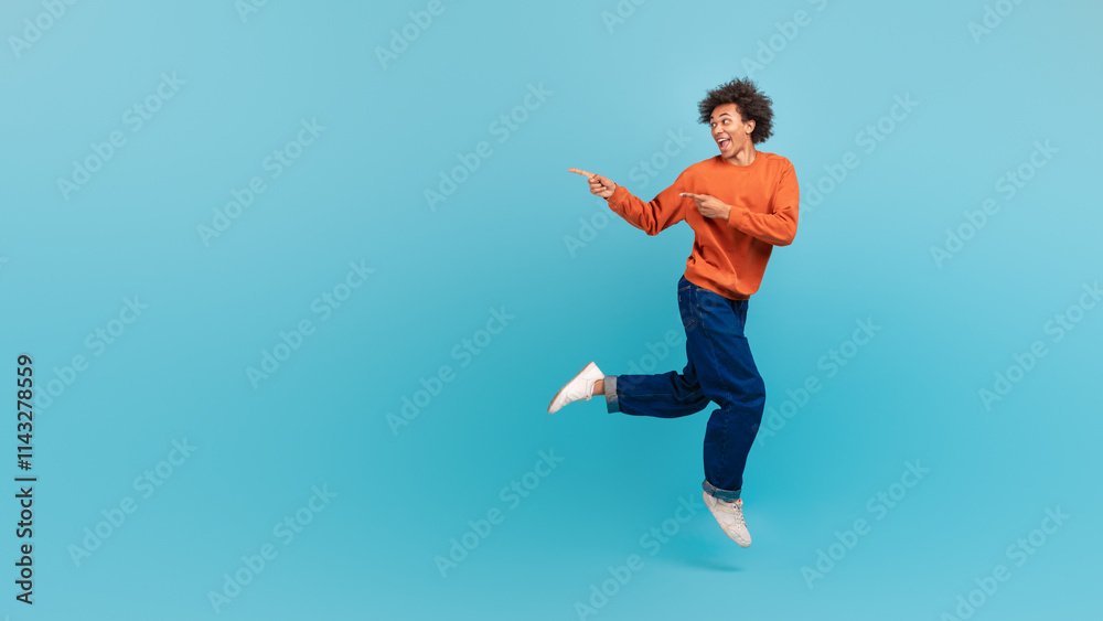Fototapeta premium A young African American man with curly hair is playfully dancing against a vibrant blue background. His energetic movements express joy and enthusiasm, capturing a fun moment, copy space