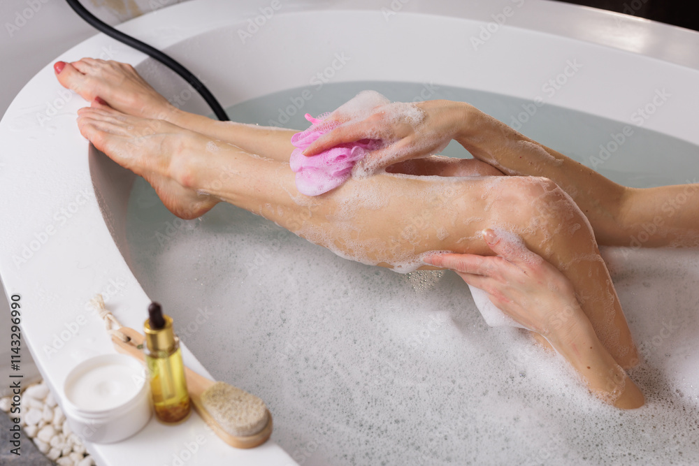 Fototapeta premium Skin care concept. Cropped view of young adult woman taking bath, lying in soap foam water, holding sponge in hand, washed leg