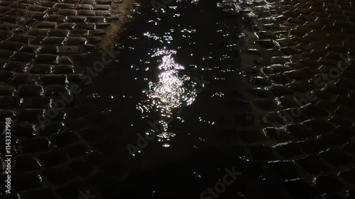 Reflection of light from a street lamp in a puddle of water during rain at night. A cobblestone street in the city.
