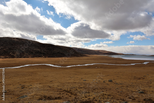 A narrow winding river crossing a flat yellowed steppe flows into a large frozen lake on an autumn cloudy day.