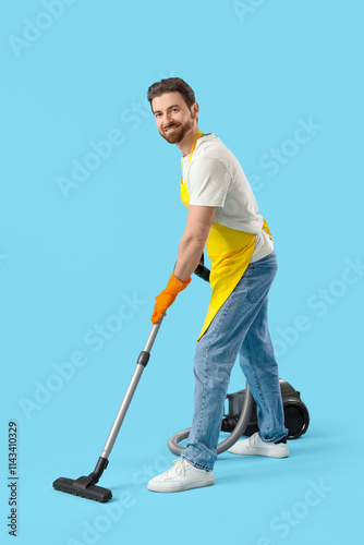 Handsome man in apron using vacuum cleaner on blue background