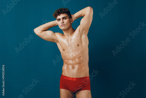 Confident young man with muscular physique posing in red swimwear against a solid teal background, showcasing strength and fitness. photo