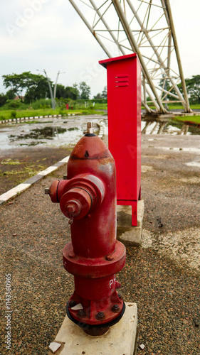 Close-up of a red fire hydrant and red utility box in an outdoor setting, with a background of greenery and industrial structures. The scene is slightly wet, indicating recent rain.