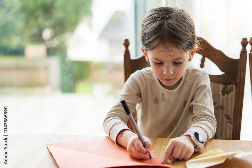 Little cute girl doing origami sitting at the table with a big window in the background. Children's education and creativity concept Little cute girl doing origami sitting at the table with a big window in the background. Children's education and creativity concept