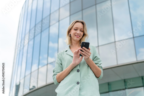 White Businesswoman Using Phone Outdoors In Front a Modern Glass Office Buildings