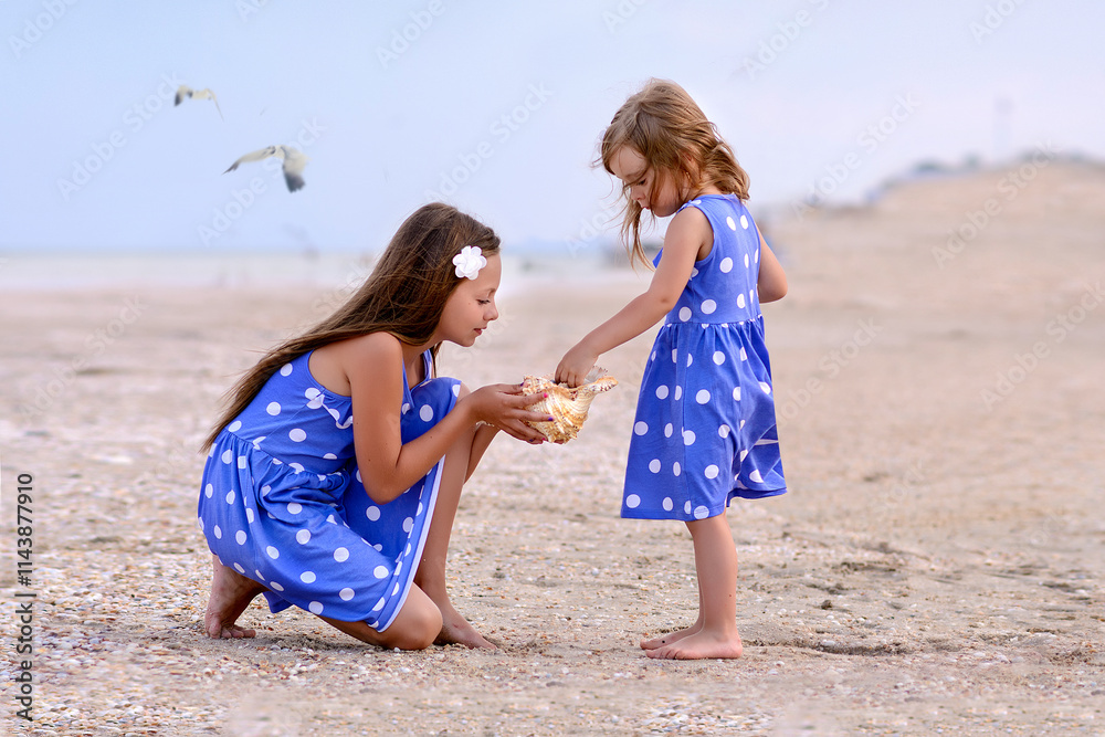 Family photo of sisters. Girls are walking on the beach near the sea, looking for a shell, smiling, hugging, they are happy and joyful. Dressed in identical dresses. Blurred background. Family photo of sisters. Girls are walking on the beach near the sea, looking for a shell, smiling, hugging, they are happy and joyful. Dressed in identical dresses. Blurred background.