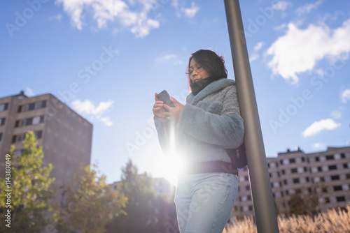 Low angle view of woman using mobile phone and leaning on pole against sky