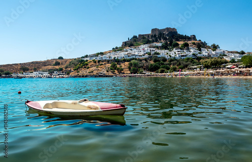 Old kayak boat in shallows of sandy beach in Bay of Lindos, with Lindos town and  Acropolis in background (Rhodes, Greece)