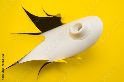 Female hand holding white clitoral vacuum vibrator sticking out of yellow paper background.  photo