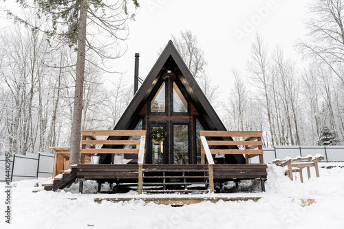 small A-frame cabin with natural wood siding, floor-to-ceiling glass panels at front. narrow wooden deck wrap around house. wooden house in snow-covered coniferous forest. Glamping in winter.