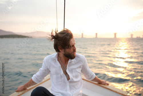 handsome man with a beard at sunset on a yacht wearing a white linen shirt and pants looking at the sea with a brutal expression