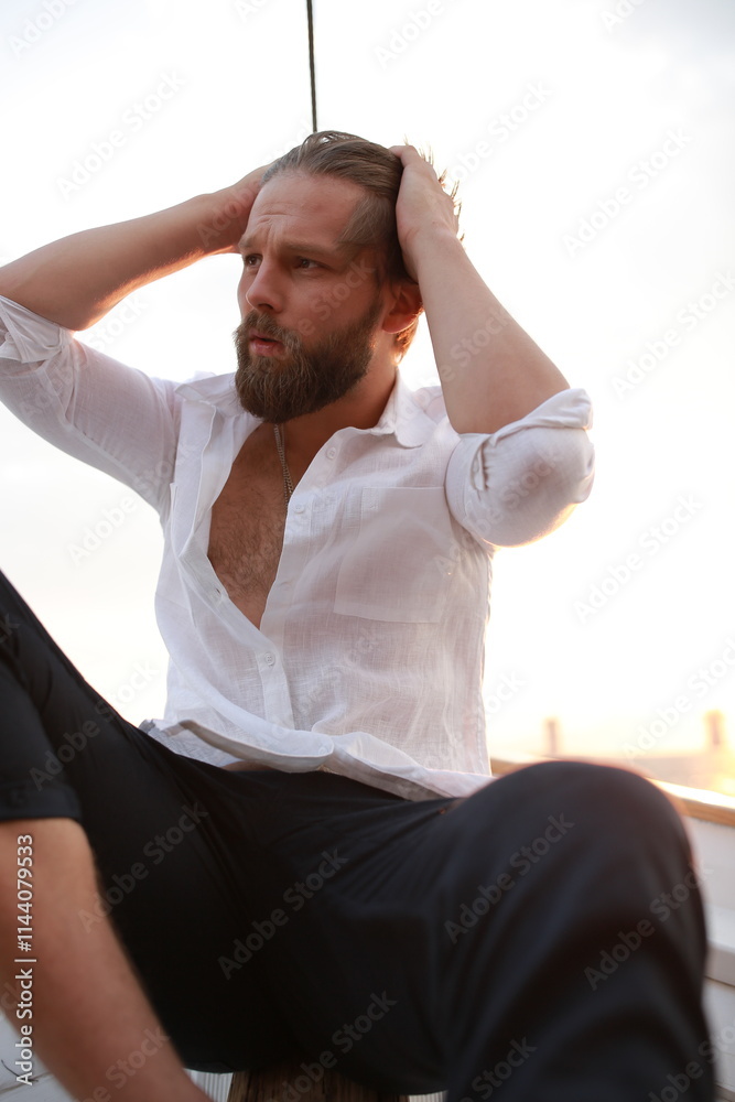 handsome man with a beard at sunset on a yacht wearing a white linen shirt and pants looking at the sea with a brutal expression handsome man with a beard at sunset on a yacht wearing a white linen shirt and pants looking at the sea with a brutal expression