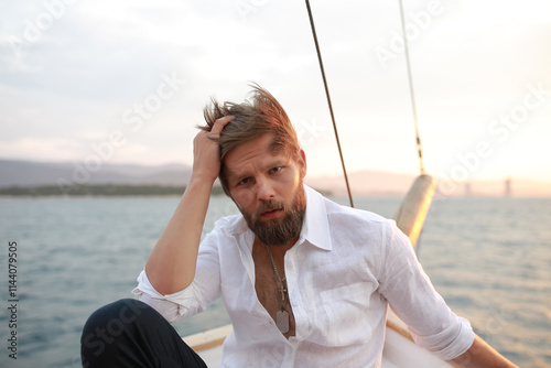 handsome man with a beard at sunset on a yacht wearing a white linen shirt and pants looking at the sea with a brutal expression