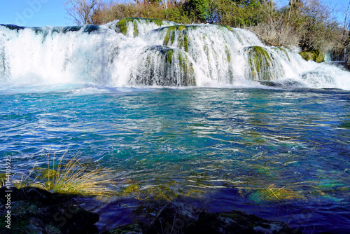 View of the Kočuša waterfall - a natural landmark in the vicinity of Ljubuski in western Bosnia and Herzegovina. A cascade of water flowing over mossy rocks and the bright blue surface of the river. photo