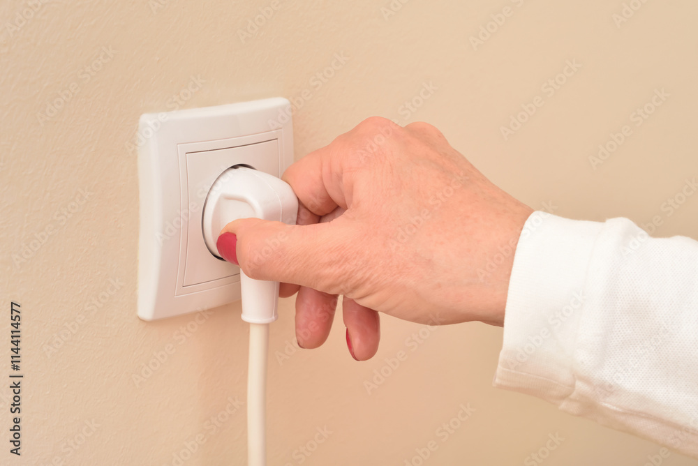 Hand of a woman holding and plugging or unplugging white electric cable in the white power socket on the pale orange wall. Copy space. Close-up. Concept of electricity consumption. Hand of a woman holding and plugging or unplugging white electric cable in the white power socket on the pale orange wall. Copy space. Close-up. Concept of electricity consumption.