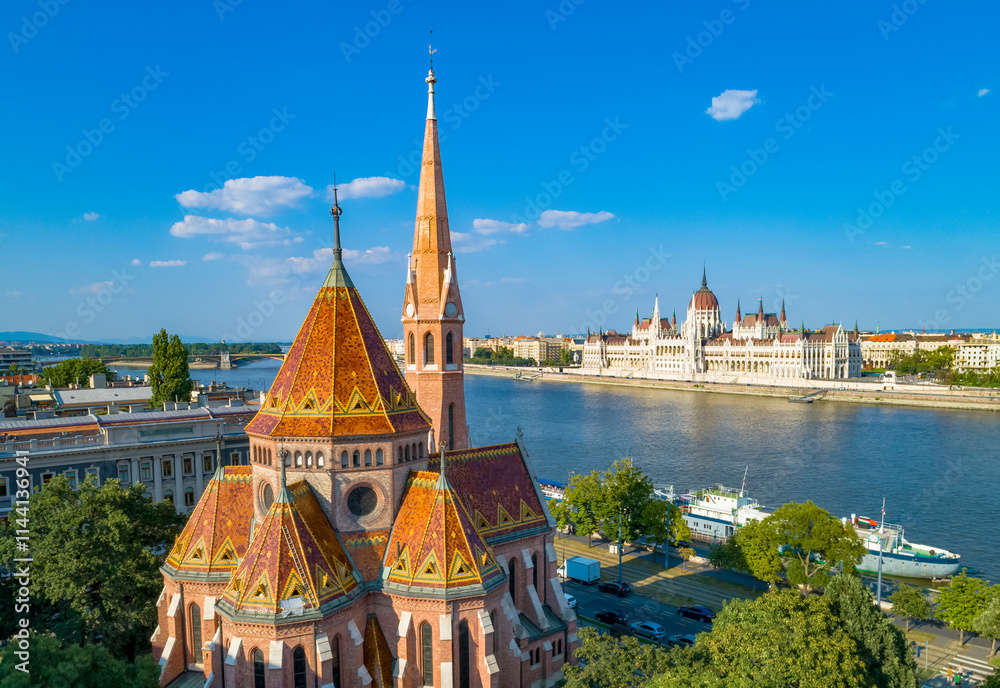 View of Budapest along the Danube River, in the foreground the Reformed Church with the patterned roof tiles and tall spire, in the background the Hungarian Parliament Building