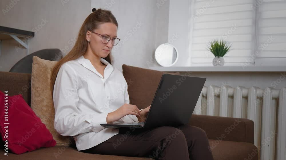 Businesswoman woman with glasses sitting on sofa in her living room and working on laptop. She wears white shirt and brown pants Businesswoman woman with glasses sitting on sofa in her living room and working on laptop. She wears white shirt and brown pants