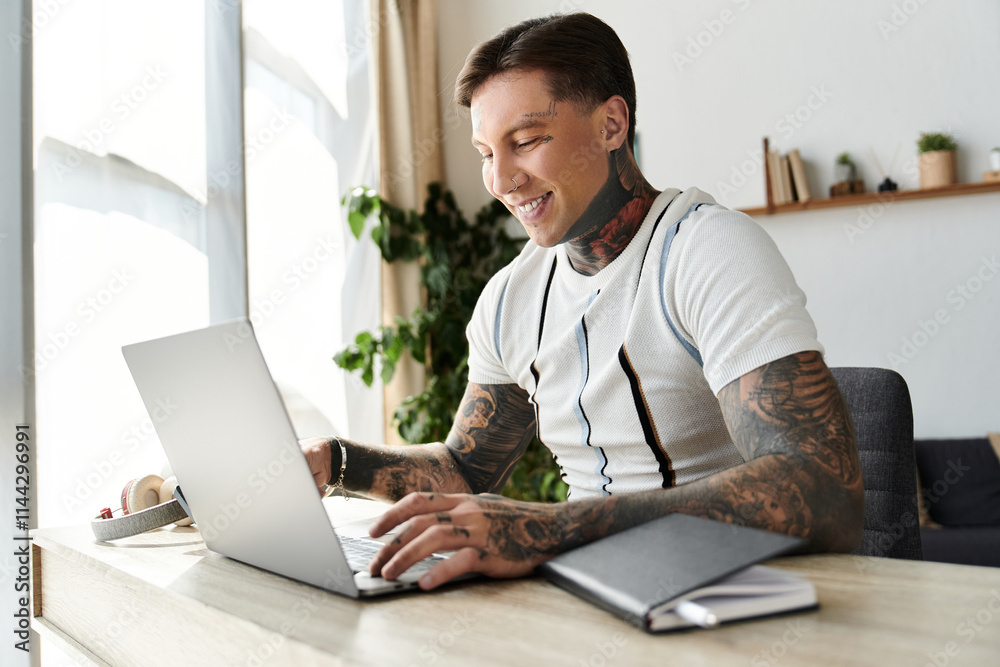 Man with tattoos sits at a desk smiling while typing on a laptop in a stylish setting. Man with tattoos sits at a desk smiling while typing on a laptop in a stylish setting.