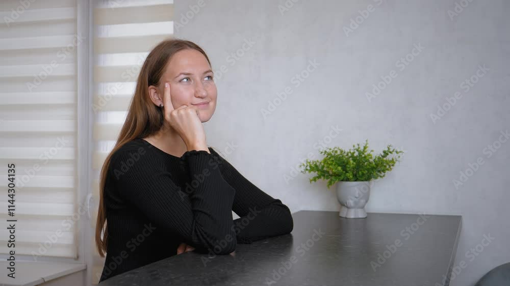 Young woman leans on countertop in modern kitchen, daydreaming while wearing black dress. Relaxation fills space as she enjoys peaceful moment of contemplation