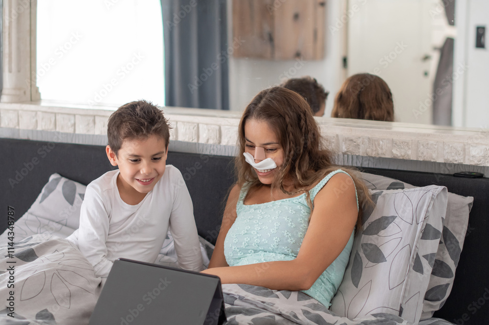 Smiling mother with bandage on nose lying in cozy bed and playing app on tablet with son, family relaxing lying among pillows on weekend, Smiling mother with bandage on nose lying in cozy bed and playing app on tablet with son, family relaxing lying among pillows on weekend,