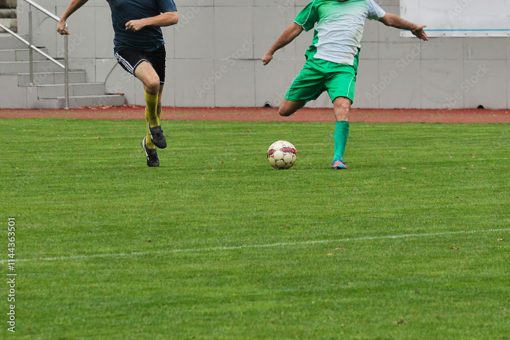 Football players in green and blue uniforms competing against each other. Dynamic photograph of a footballer in motion, preparing to strike the ball on the football field.