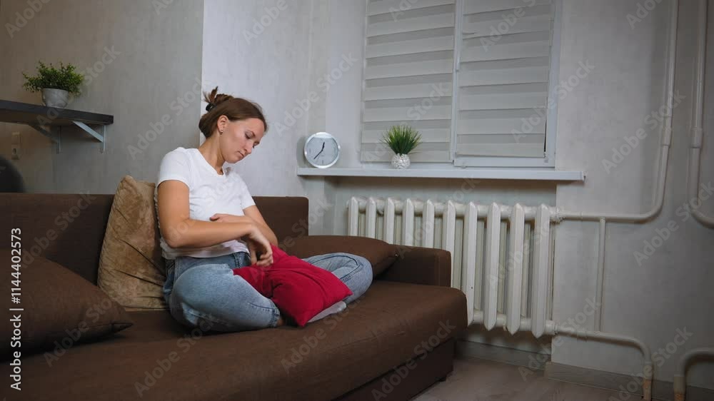 Young woman sitting on sofa, clutching her stomach in pain, shows signs of discomfort and distress. Expressions of worry and anxiety reflect her struggle with abdominal issues Young woman sitting on sofa, clutching her stomach in pain, shows signs of discomfort and distress. Expressions of worry and anxiety reflect her struggle with abdominal issues