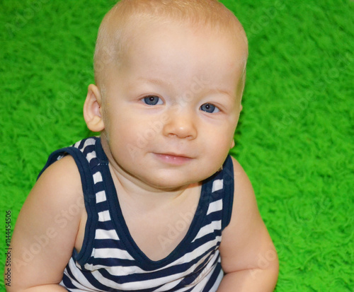 A cute 6-month-old baby in a striped T-shirt on a green background looks directly at the camera