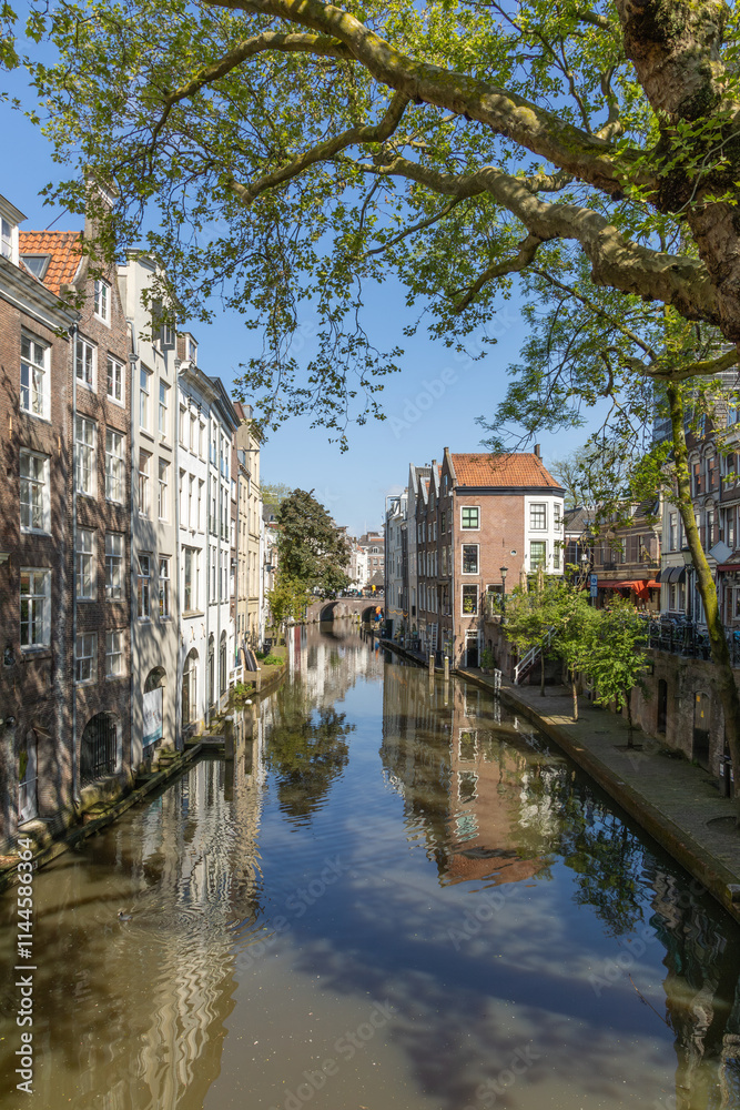 Fototapeta premium Tall Canal houses along the Oudegracht canal in Utrecht with a wharf and a bridge in the distance. A blue sky.