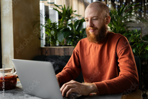 Focused red-haired bearded millennial man sitting at table in cafe. serious freelance businessman working or studying on computer remotely looks with smile